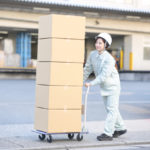 屋外でダンボール箱を台車に載せて運ぶ作業服の女性