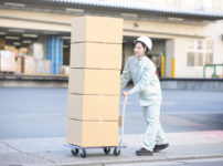 屋外でダンボール箱を台車に載せて運ぶ作業服の女性