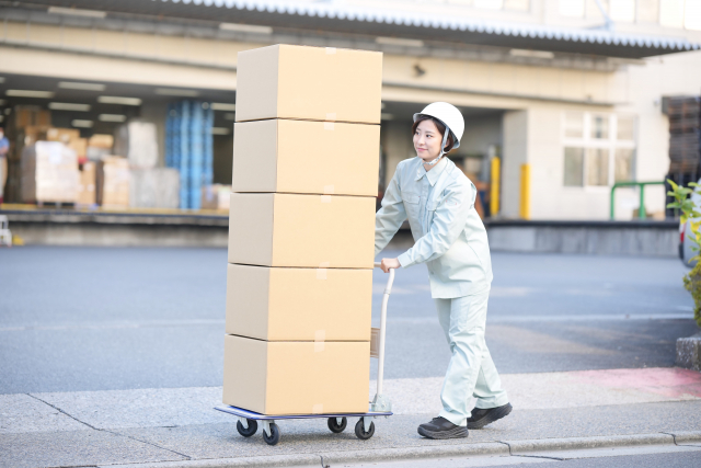 屋外でダンボール箱を台車に載せて運ぶ作業服の女性